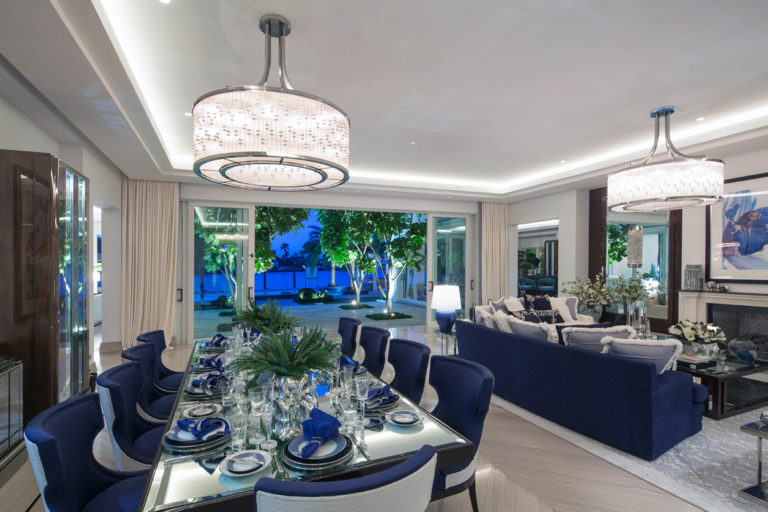 A residential living room lit with LED linear strip cove lighting and large drumb chandeliers, open to an outdoor courtyard at dusk. Boca Lighting and Controls