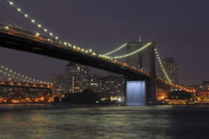 The Brooklyn Bridge lit in early evening with the cityscape in the distance. Under the bridge, LED lighting illuminates a featured waterfall. Boca Lighting and Controls