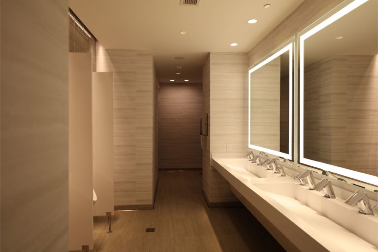 Linear LED strip lights illuminating the edges of large mirrors in the vanity area of a modern hotel public restroom. Boca Lighting and Controls