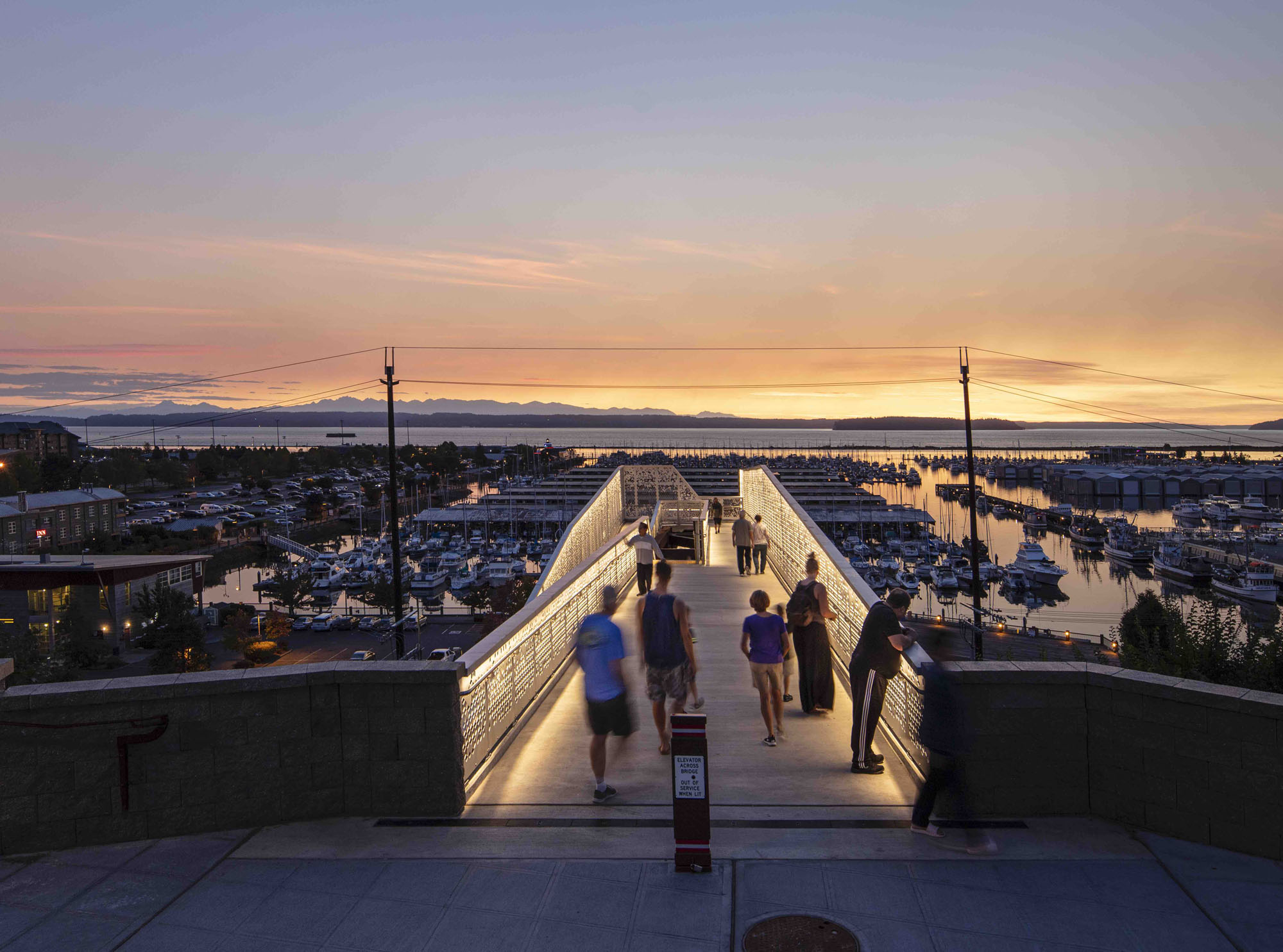 LED strip lights illuminating architectural steel and pathways on a pedestrian bridge overlooking a marina at sunset. Boca Lighting and Controls